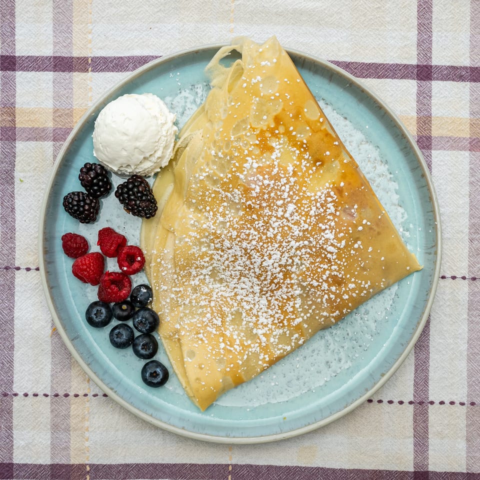 Golden crepes dusted with powdered sugar, served with whipped cream and mixed berries on a blue plate.