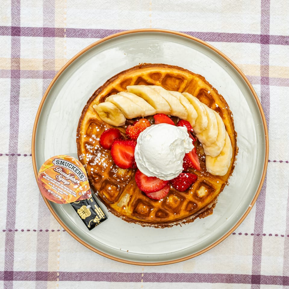 Golden waffle topped with sliced bananas, strawberries, whipped cream, and powdered sugar on a plate with syrup and butter.