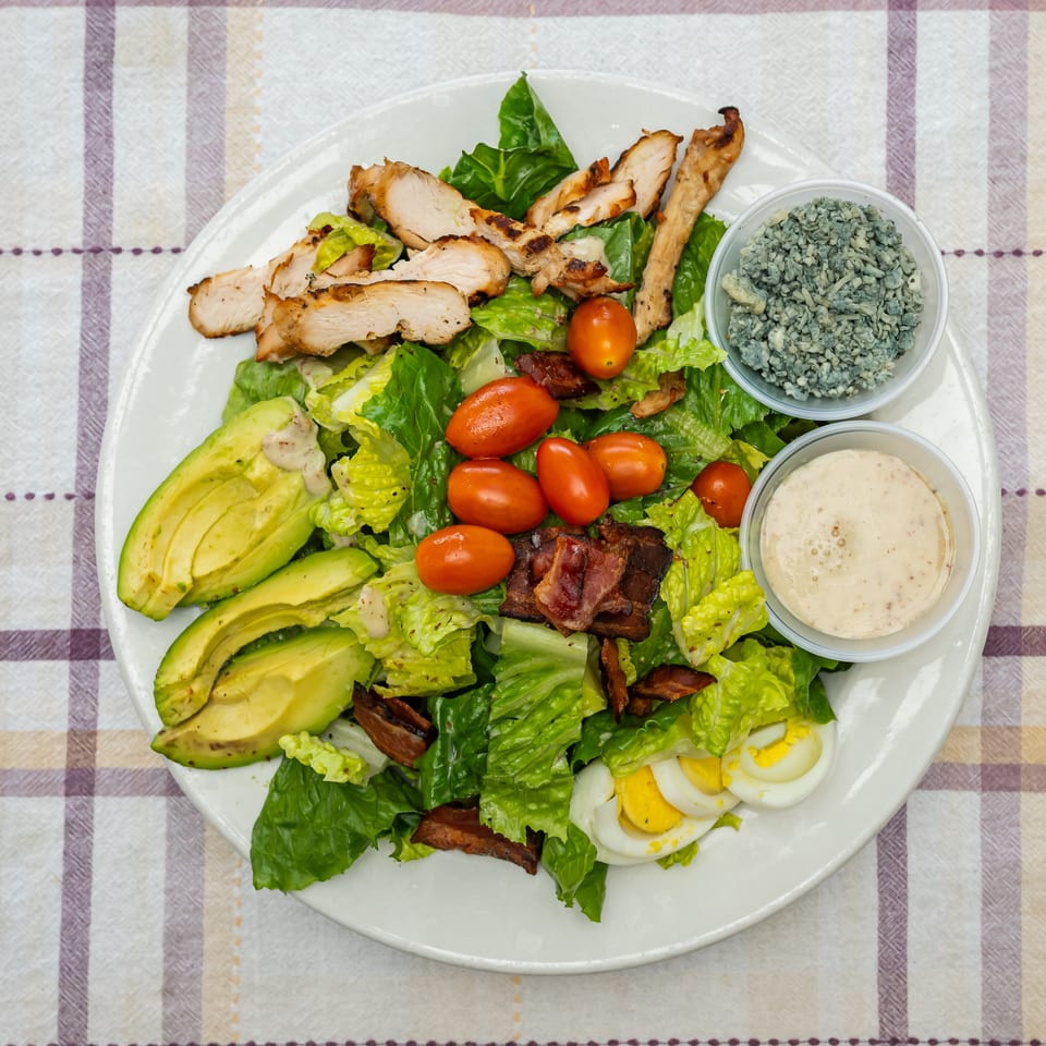 Grilled chicken Cobb salad with romaine, avocado, tomatoes, bacon, egg, blue cheese, and dressing cups.