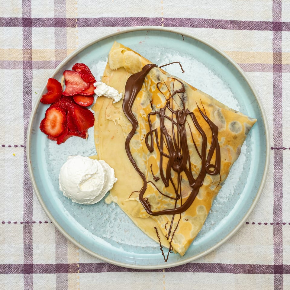 Folded crepes drizzled with chocolate, served with sliced strawberries, whipped cream, and vanilla ice cream on a blue plate.