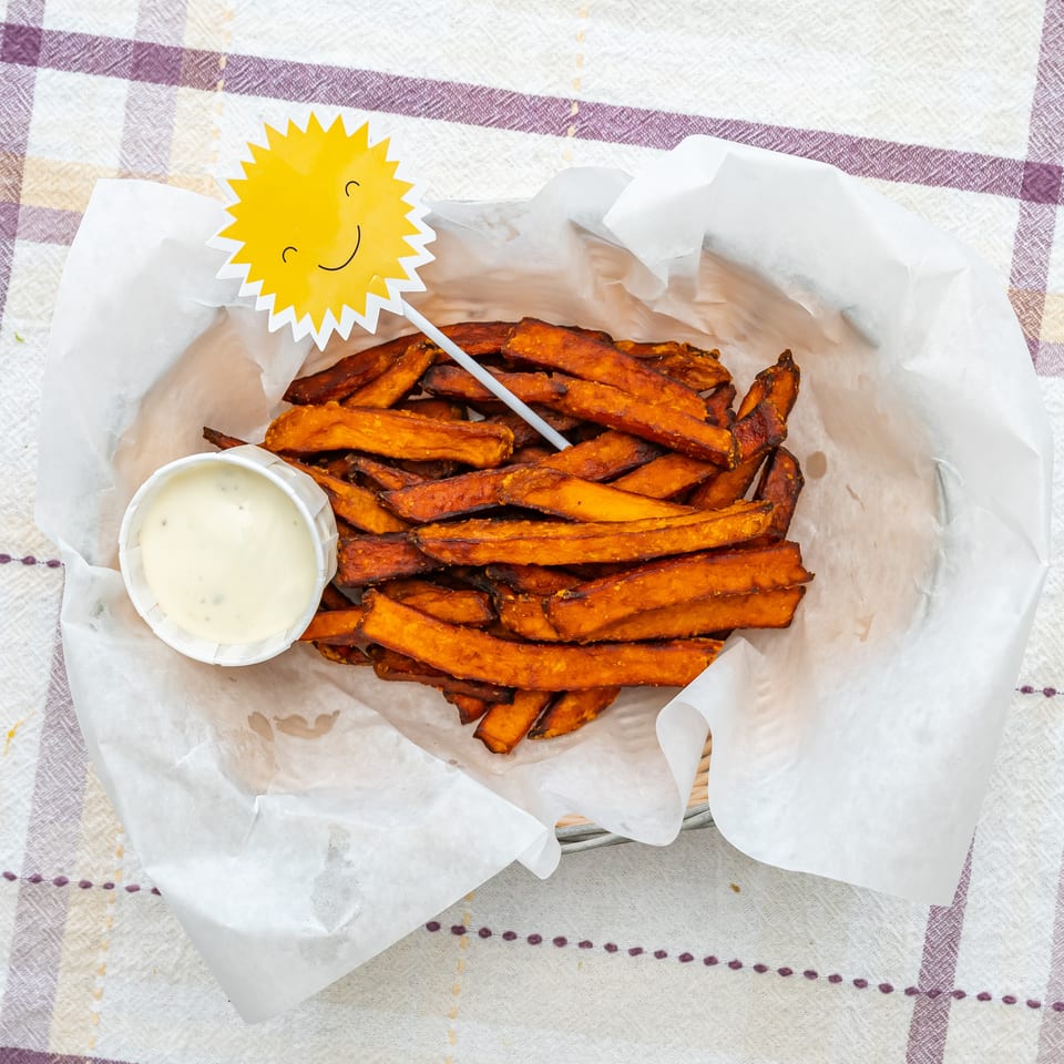 Crispy sweet potato fries served in parchment with a cup of creamy dipping sauce on a checkered tablecloth.