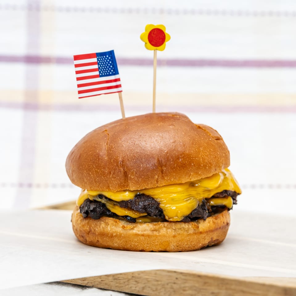 Double cheeseburger on a toasted bun, topped with American flag and sunflower decorations, against a softly patterned background.