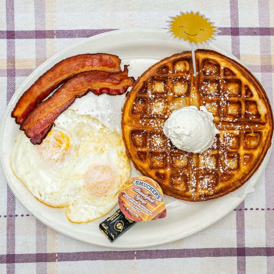 Breakfast plate with Belgian waffle, whipped cream, powdered sugar, bacon, fried eggs, syrup, and butter on a checkered tablecloth.